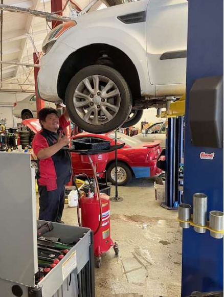 Inside Nextgen Auto Center service bay with a mechanic dressed in red and blue working on a white car that is on a lift. In the background, you can see a red car with the hood up and other vehicles